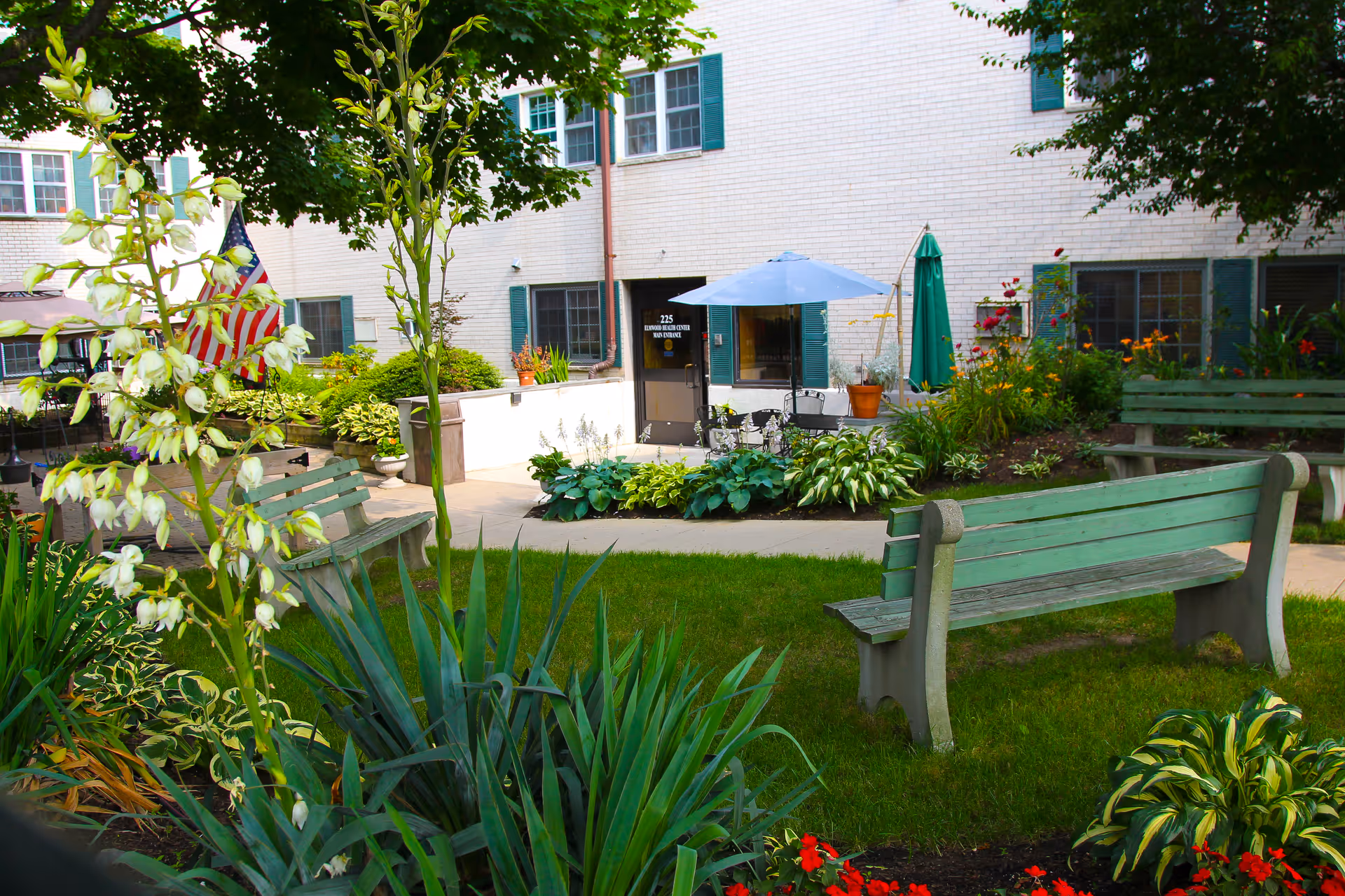 Outdoor garden area at Elmwood Nursing & Rehabilitation Center featuring green benches, various plants and flowers, a small patio with a table and umbrella, and the building's white brick exterior with green window shutters.