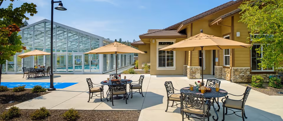 Outdoor patio area at Eskaton Village Placerville with several round metal tables and chairs under large beige umbrellas. The patio is paved with concrete and surrounded by landscaped plants. In the background, there is a large glass greenhouse structure and a building with beige siding and stone accents.