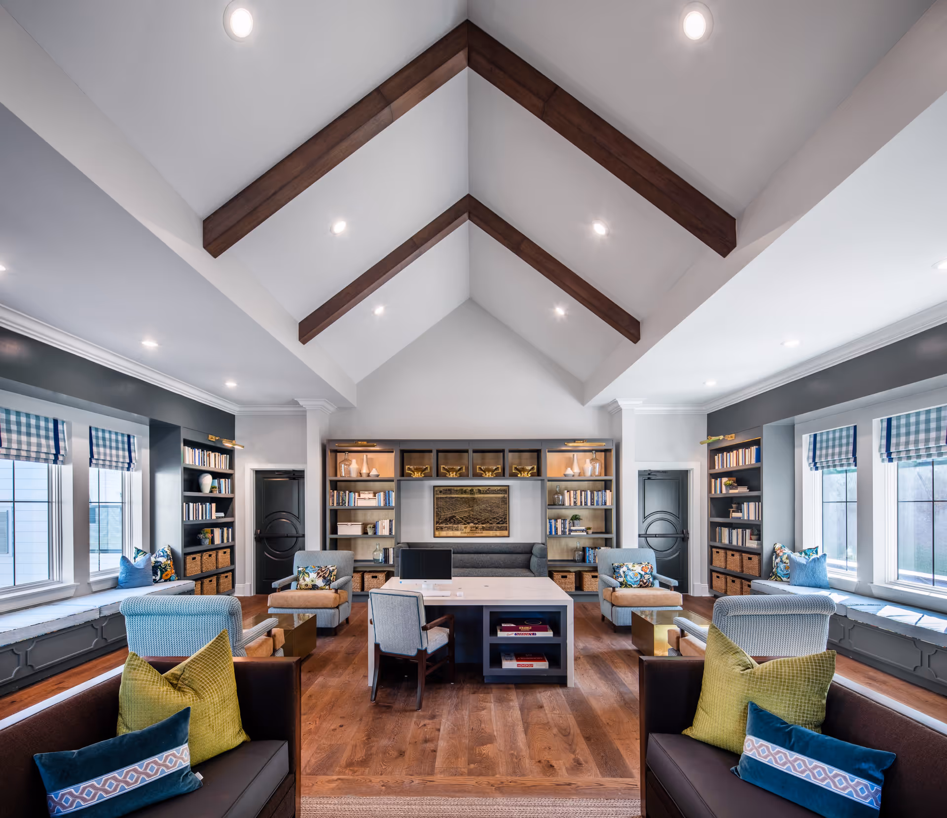 A spacious and well-lit living room with a vaulted ceiling featuring exposed wooden beams. The room has large windows with blue and white checkered valances, built-in bookshelves filled with books and decorative items, and comfortable seating including armchairs and sofas with colorful cushions. A central table with chairs is placed on a wooden floor, creating a cozy and inviting atmosphere.