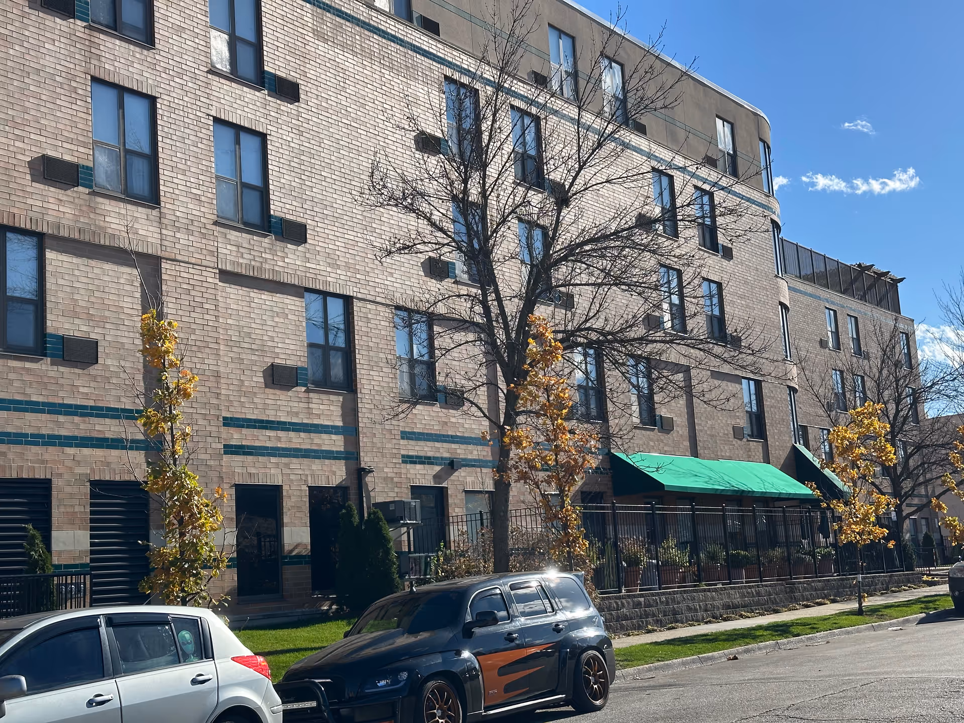 Exterior view of a multi-story brick building with several windows, small trees with autumn leaves in front, a green awning over an entrance, and parked cars along the street under a clear blue sky.
