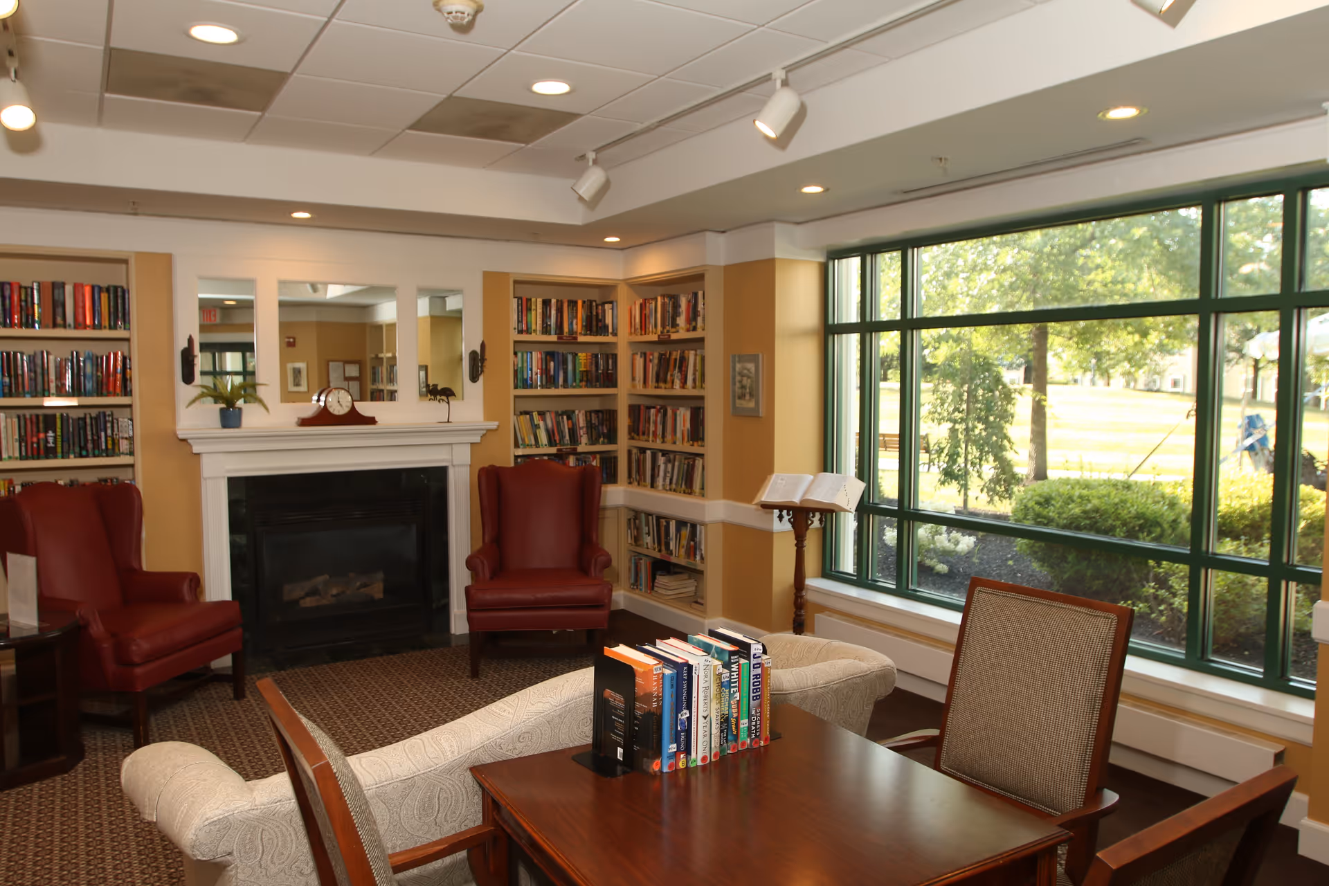 A cozy reading room with large windows overlooking a green outdoor area. The room features two red leather armchairs, a white patterned sofa, a wooden table with books standing upright, built-in bookshelves filled with books, a fireplace with a clock on the mantel, and a music stand holding an open book.