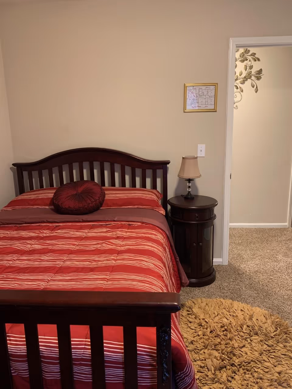 A bedroom with a dark wooden bed frame and a red striped comforter with a round red pillow. Next to the bed is a round dark wooden nightstand with a small lamp. The room has beige walls and carpeted floor with a round beige rug. There is an open doorway showing part of another room with a decorative wall decal.