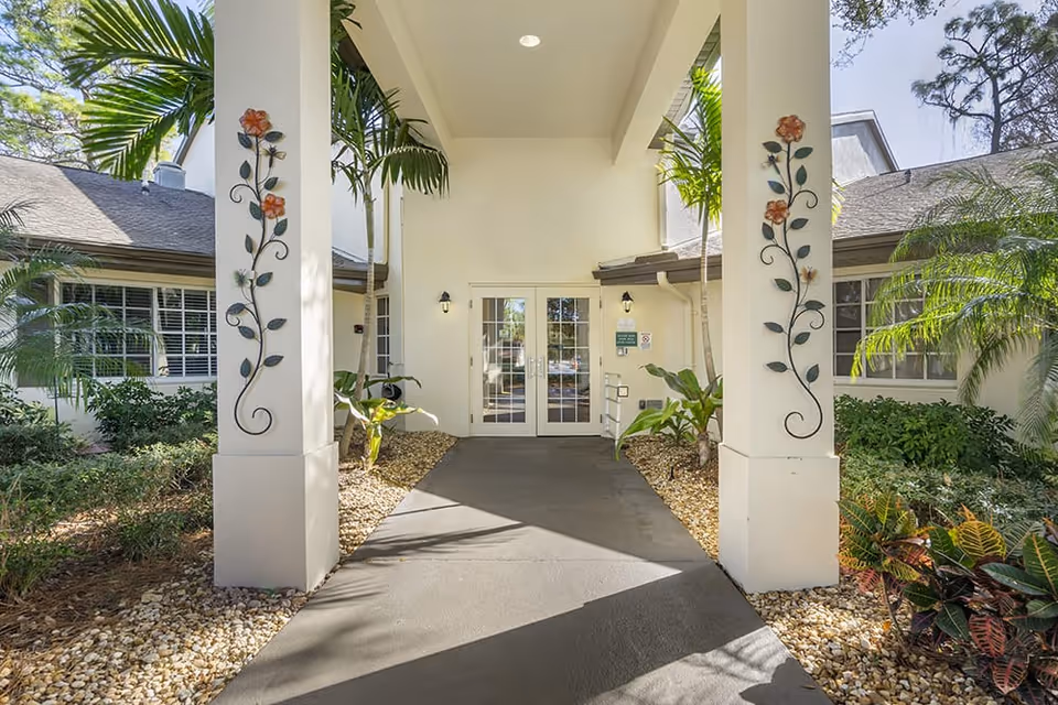 Entrance to a building with two white pillars decorated with metal floral art. The pathway is surrounded by small rocks and tropical plants, leading to double glass doors under a covered porch.