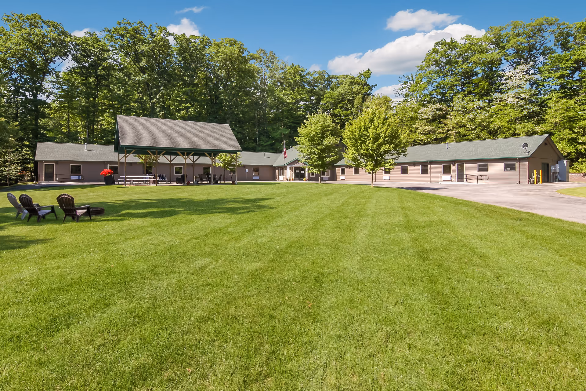 Wide view of South Torch Assisted Living facility showing a large green lawn with a few chairs and a fire pit on the left. The building is a single-story structure with a covered outdoor seating area in the center, surrounded by trees under a blue sky with some clouds.