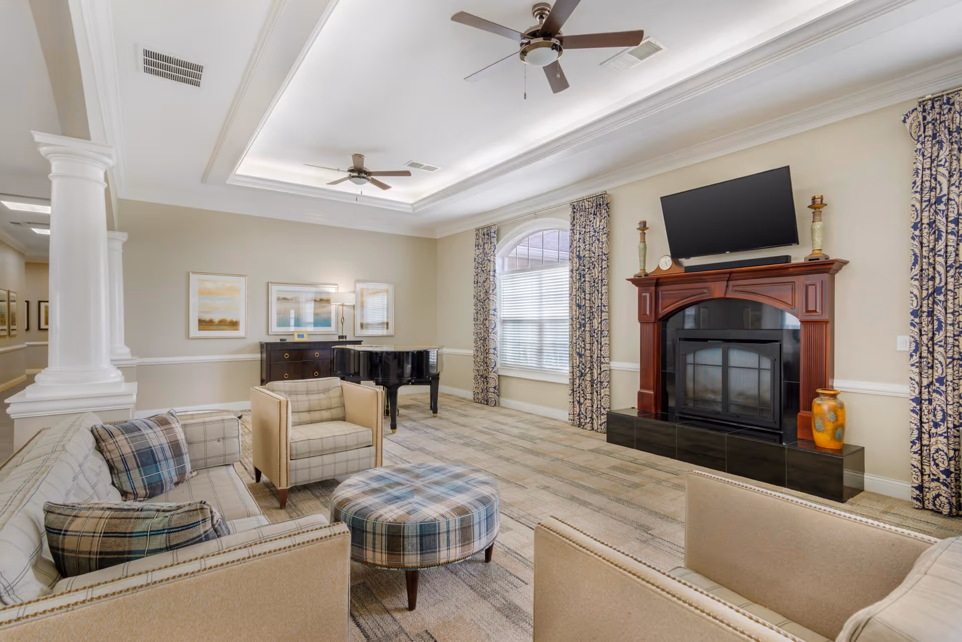 Spacious lounge with sofas, armchairs, a plaid ottoman, a grand piano, and a fireplace topped by a wall-mounted TV.