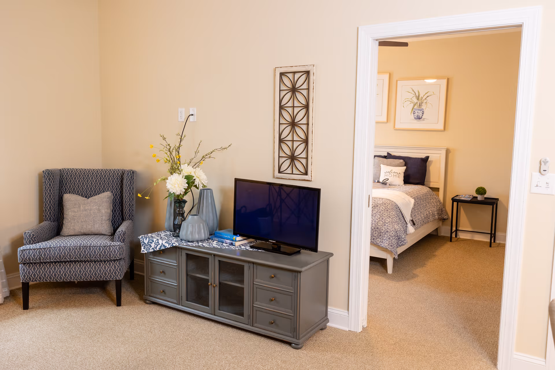 A cozy living room area with a patterned armchair, a gray TV stand holding a flat-screen television, and decorative vases with flowers. Through an open doorway, a bedroom is visible with a bed featuring blue and white bedding, a 'welcome home' pillow, framed botanical artwork on the wall, and a small black side table with a plant and books.