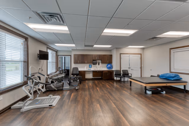 A spacious room with exercise equipment including a recumbent bike and elliptical machine, a treatment table with a blue pillow, two chairs, and a kitchenette area with cabinets, sink, and countertop. The room has wood flooring, white walls, and several windows with blinds allowing natural light.
