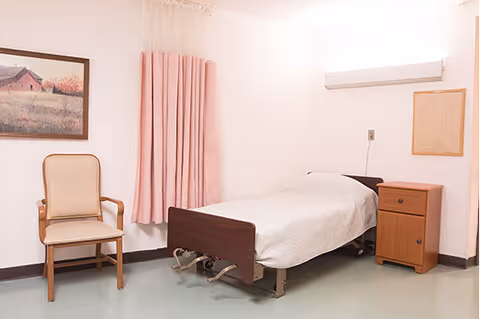A simple bedroom with a single hospital-style bed covered with a white blanket, a wooden nightstand with two drawers, a beige upholstered chair with wooden arms, a framed painting of a barn on the wall, and pink curtains covering a window.