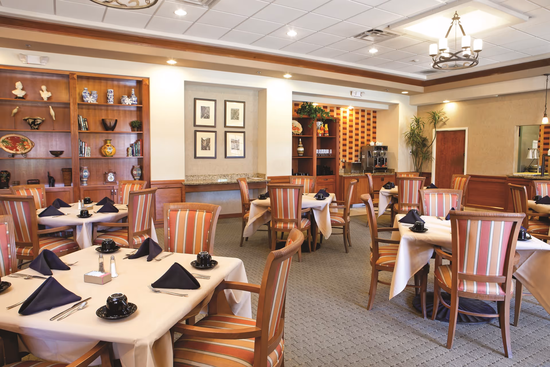 A dining room with multiple tables covered in beige tablecloths, each set with black napkins, black cups and saucers, and silverware. The chairs have wooden frames with striped upholstery in shades of red, orange, and beige. The room features built-in wooden shelves with decorative items and framed pictures on the walls. There is a coffee station in the back corner and a chandelier hanging from the ceiling.