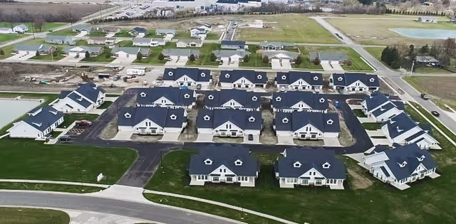 Aerial view of a residential community with multiple single-story houses featuring dark roofs and white walls, arranged in neat rows with paved roads and green lawns surrounding them.