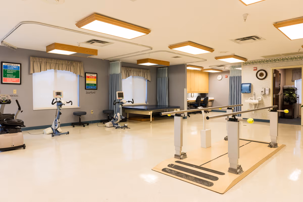 A rehabilitation room with exercise bikes, parallel bars for walking practice, a treatment table, and a desk area with chairs. The room has large windows with curtains, bright overhead lighting, and medical posters on the walls.