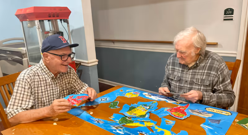 Two elderly men sitting at a wooden table playing a puzzle game with colorful pieces. One man is wearing a plaid shirt and a baseball cap, smiling, while the other man, also in a plaid shirt, is focused on the puzzle pieces. Behind them is a popcorn machine and a wall with a room number sign 209.
