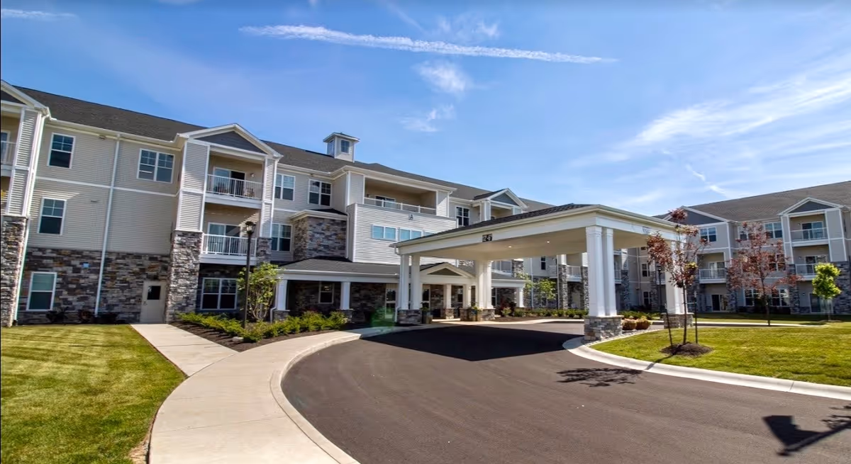 Exterior view of a modern three-story senior living facility with beige siding and stone accents, featuring balconies, a covered entrance driveway, well-maintained landscaping, and a clear blue sky.