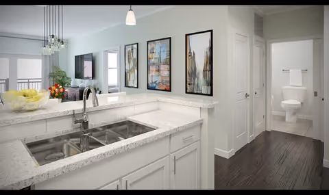 Open kitchen with a double sink and white speckled countertops opening to a living area with framed artwork and a hallway leading to a bathroom with a visible toilet.
