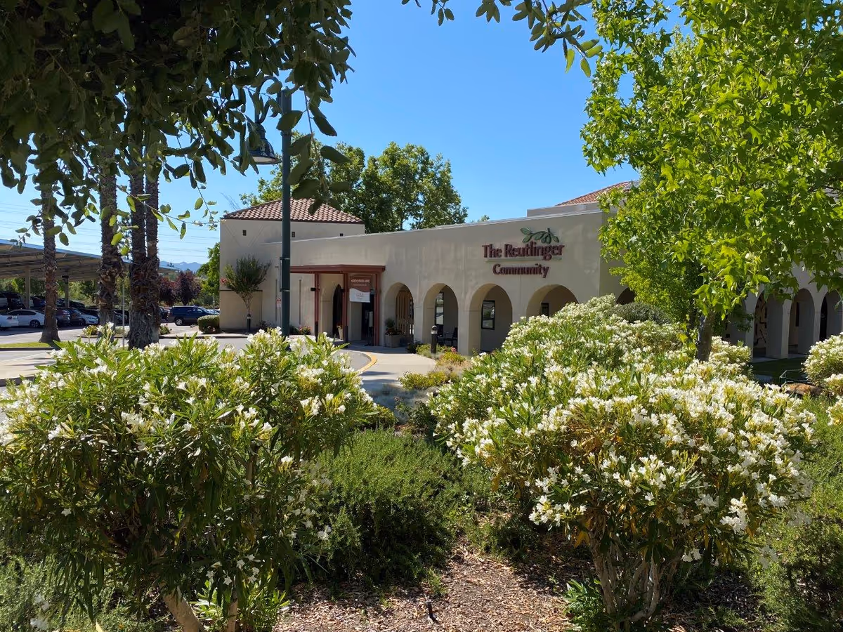 Exterior view of The Reutlinger Community building with arched entrances, surrounded by green trees and bushes with white flowers under a clear blue sky.