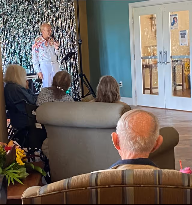 An elderly man and several other seniors are seated in a common area watching a man in a colorful shirt perform with a microphone in front of a shiny silver curtain backdrop.