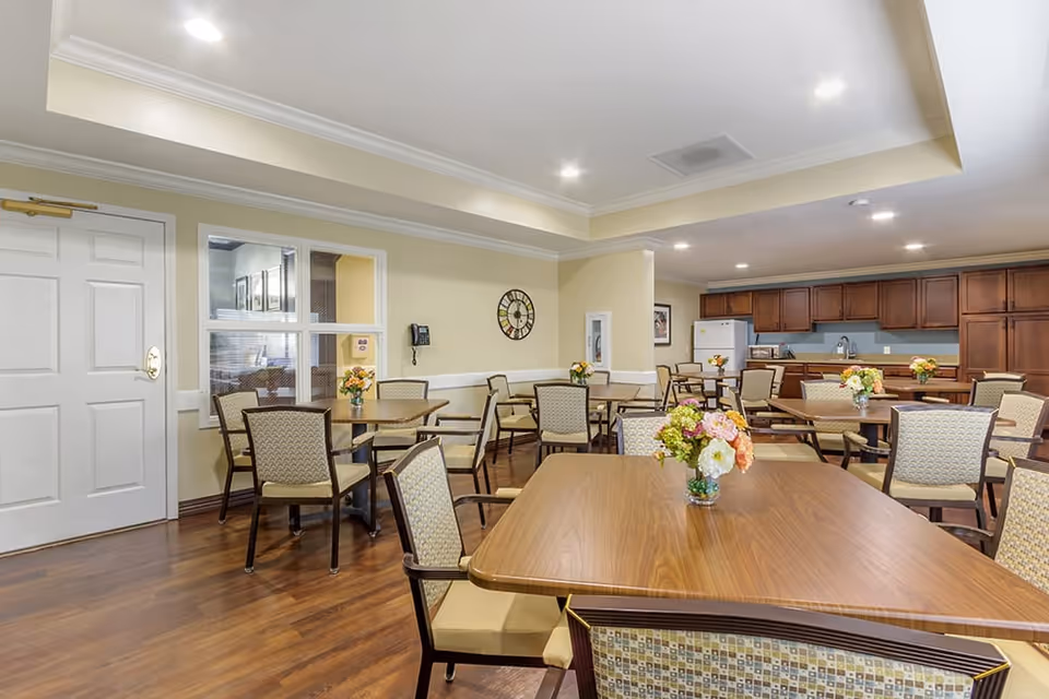 A well-lit dining room with multiple wooden tables and cushioned chairs arranged neatly. Each table has a small vase with colorful flowers. The room features wooden flooring, cream-colored walls, a clock on the wall, and a kitchen area with wooden cabinets and a white refrigerator in the background.