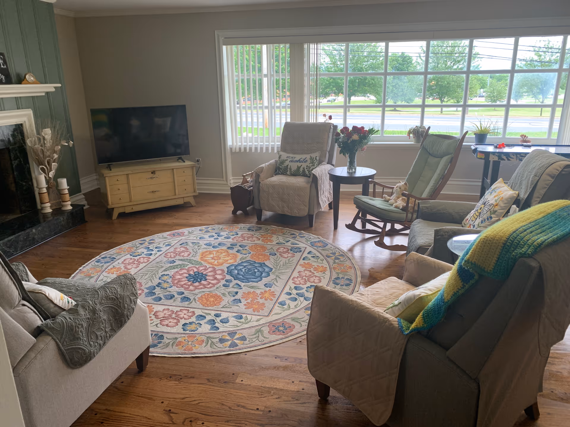 A cozy living room with a large window showing a view of trees and a road outside. The room features a round floral patterned rug on wooden floors, several cushioned armchairs and a rocking chair arranged around a small round table with a vase of flowers. A flat-screen TV sits on a light-colored wooden cabinet near a fireplace with decorative candles and dried plants.