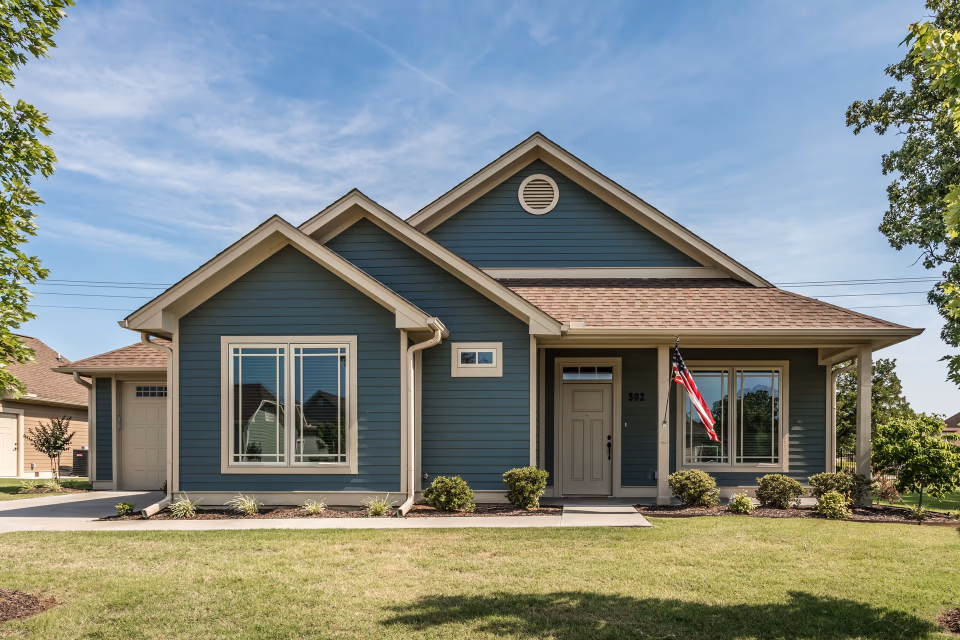 Front exterior view of a single-story blue house with beige trim, a small front porch with an American flag, multiple windows, a garage on the left side, and a well-maintained lawn with shrubs.