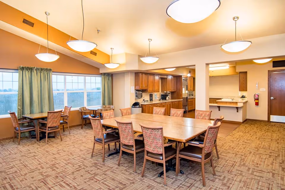 A well-lit dining room with a large rectangular table surrounded by chairs with patterned upholstery. There are additional smaller tables and chairs near large windows with green curtains. The room features warm beige walls, carpeted flooring, and ceiling lights. In the background, there is a kitchen area with wooden cabinets and appliances.