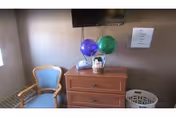 Small interior room with a wooden dresser topped by two balloons and toiletries, a blue upholstered chair, a wall-mounted TV, and a laundry basket.
