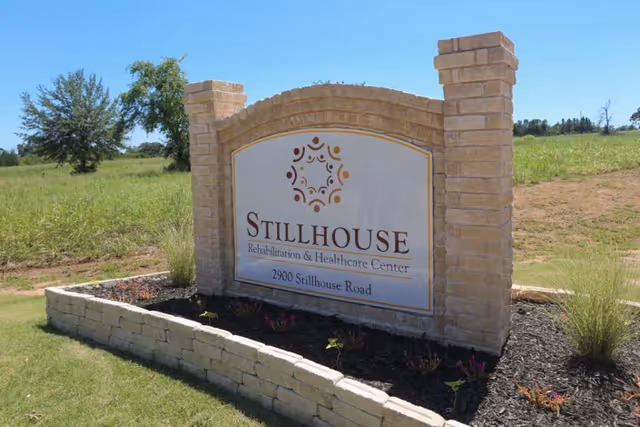 A stone sign for Stillhouse Rehabilitation & Healthcare Center located at 2900 Stillhouse Road, set in a landscaped area with grass, small plants, and a tree in the background under a clear blue sky.