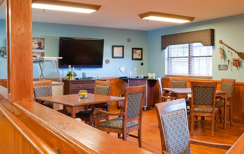 A cozy dining area with wooden tables and cushioned chairs arranged neatly. The walls are painted light blue with wooden paneling on the lower half. A flat-screen TV is mounted on the wall above a dark wooden cabinet. There are framed pictures and decorative items on the walls, and a window with blinds and a valance lets in natural light. The floor is wooden, and the room has a warm, inviting atmosphere.