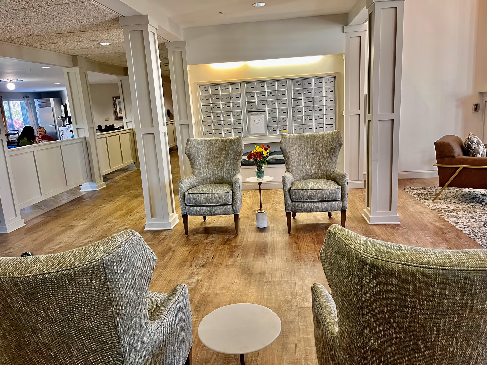 A cozy senior living facility common area with four upholstered armchairs arranged around a small round table with a vase of flowers. The room features wooden flooring, white paneled columns, and a wall of mailboxes in the background. There is a seating area with a brown leather couch and a patterned rug to the right, and a hallway with people visible in the distance to the left.