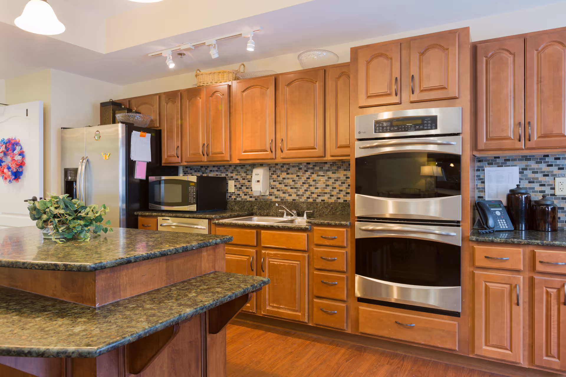 A modern kitchen with wooden cabinets, a double oven, a microwave, a stainless steel refrigerator, a sink, and granite countertops. There is a small plant on the kitchen island and a colorful wreath hanging on a white door in the background.