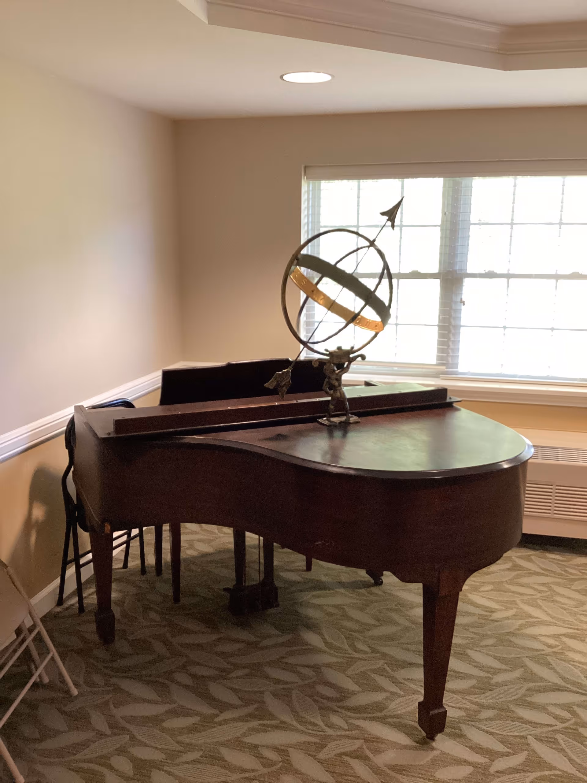 A grand piano with a decorative armillary sphere on top is placed in a carpeted room with beige walls and a large window with blinds. There are a few folding chairs stacked against the wall in the corner.