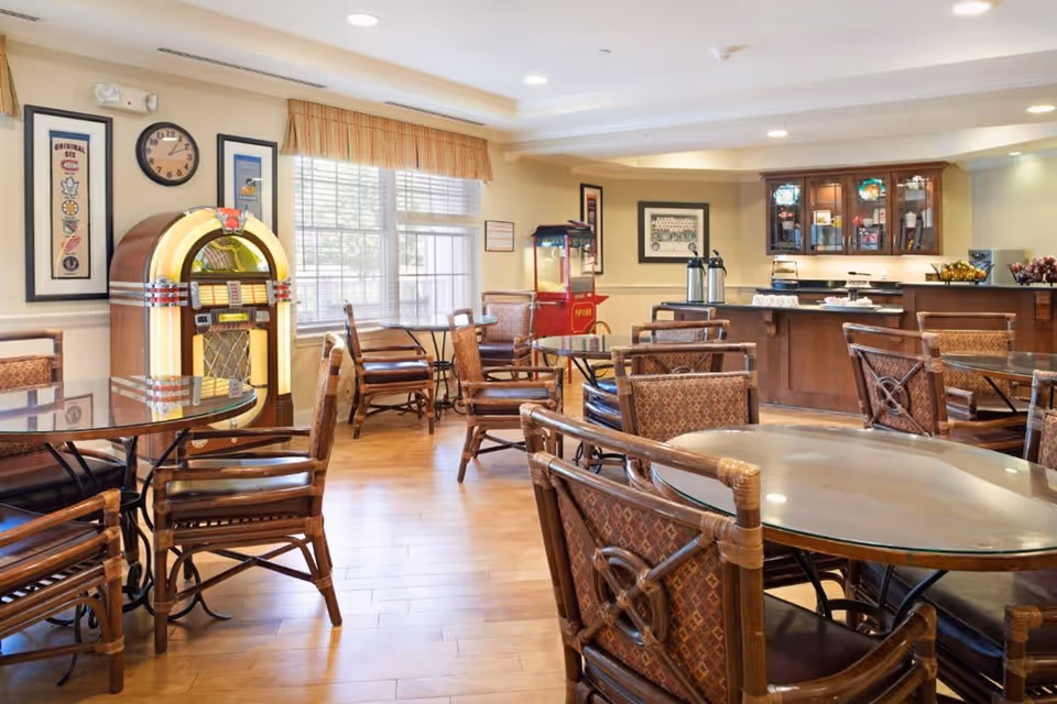 A bright and inviting common dining area with multiple round glass-top tables and wooden chairs with patterned cushions. A vintage jukebox is positioned near a window with blinds and a valance. The room features hardwood floors, framed artwork on the walls, and a counter area with coffee dispensers and a popcorn machine in the background.