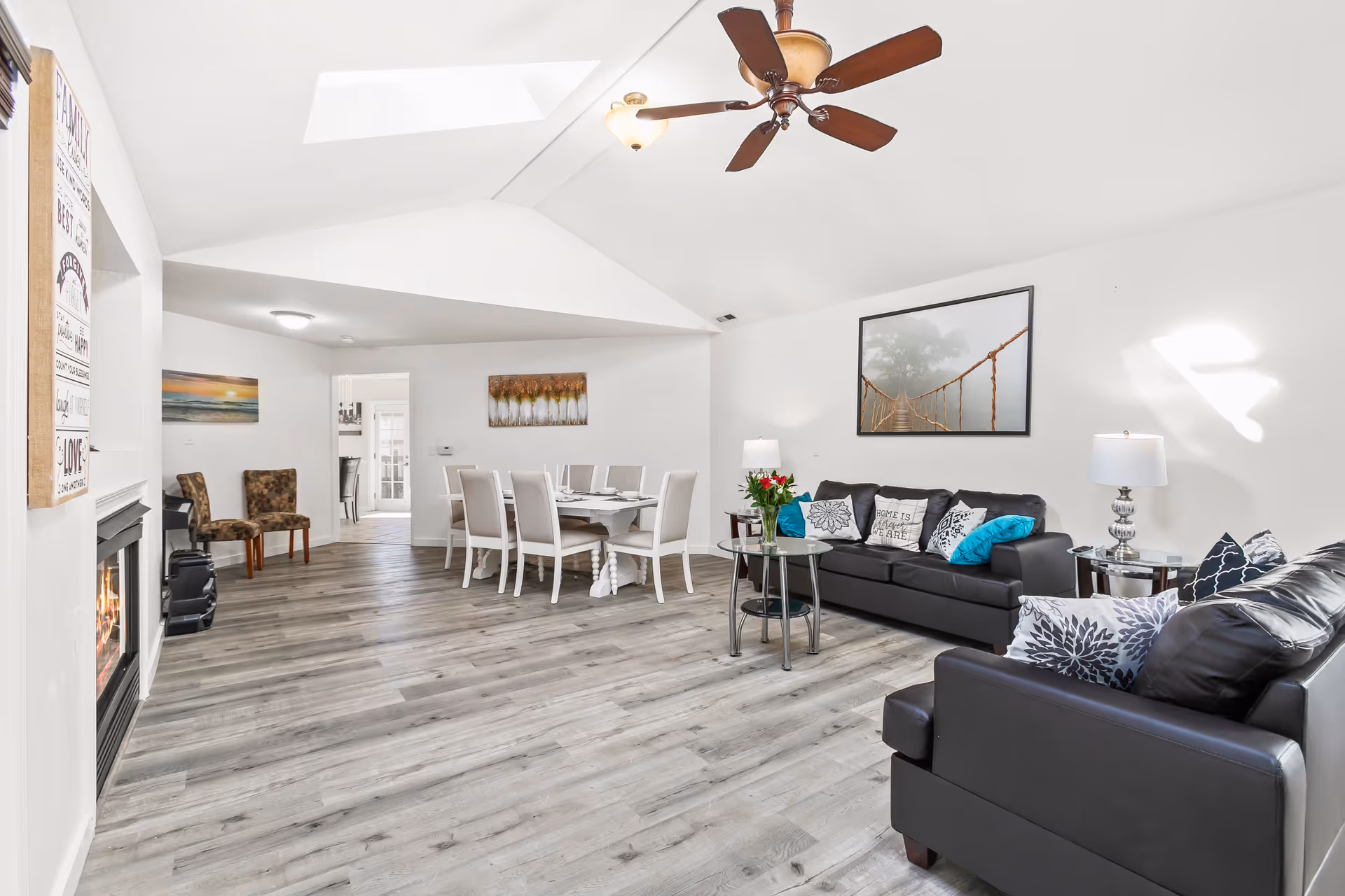 A bright and spacious living room and dining area with light gray wood flooring and white walls. The living room features two black leather sofas adorned with decorative pillows, a glass coffee table with a vase of flowers, and two side tables with lamps. A large framed picture of a foggy suspension bridge hangs on the wall. The dining area has a white dining table with six chairs. Ceiling fan with lights is mounted on the vaulted ceiling. A fireplace is visible on the left side with a decorative sign above it. Two patterned chairs are placed near the dining area.