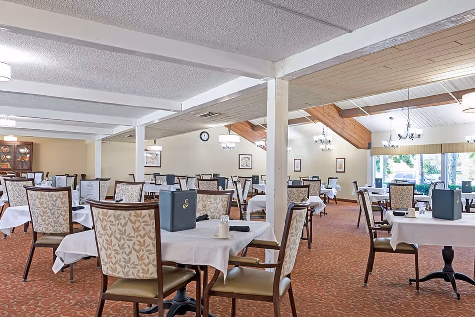 A spacious dining room with multiple tables covered in white tablecloths, each set with menus, napkins, and salt and pepper shakers. The room features patterned chairs, a carpeted floor, chandeliers, and large windows letting in natural light.
