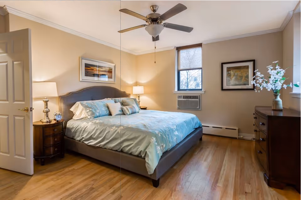 A neatly made bed with light blue floral bedding in a bedroom featuring wooden floors, a ceiling fan, two bedside tables with lamps, a window with a beige roller shade, an air conditioning unit, a wooden dresser with a vase of white flowers, and framed artwork on the walls.