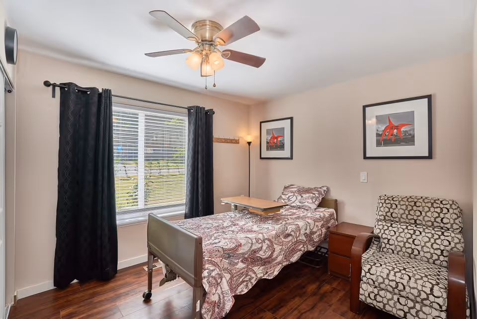 A small bedroom with a single bed covered in a patterned bedspread, a wooden overbed table, a patterned armchair, two framed pictures on the wall, a floor lamp, a ceiling fan with lights, and a window with black curtains and white blinds.