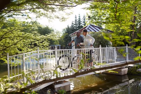 An elderly couple standing on a white metal bridge over a pond in a lush green garden. The man is holding a bicycle while the woman stands beside him. Trees and shrubs surround the area, and there is a small gazebo in the background.