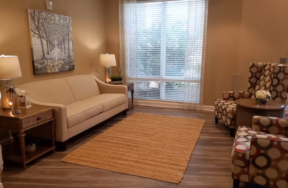 A cozy living room with a beige sofa, two patterned armchairs, a wooden side table with a lamp and decorative items, a round wooden coffee table with a small flower arrangement, a large window with sheer curtains, and a textured area rug on a wooden floor.