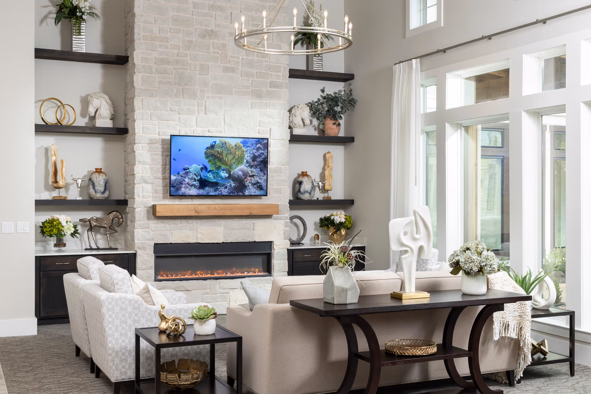 A bright and elegant living room with large windows allowing natural light to fill the space. The room features a stone fireplace with a mounted flat-screen TV above it, flanked by dark wooden shelves decorated with various sculptures, plants, and vases. There are two light-colored sofas arranged around a dark wooden console table adorned with decorative items and plants. The overall decor is modern and inviting.