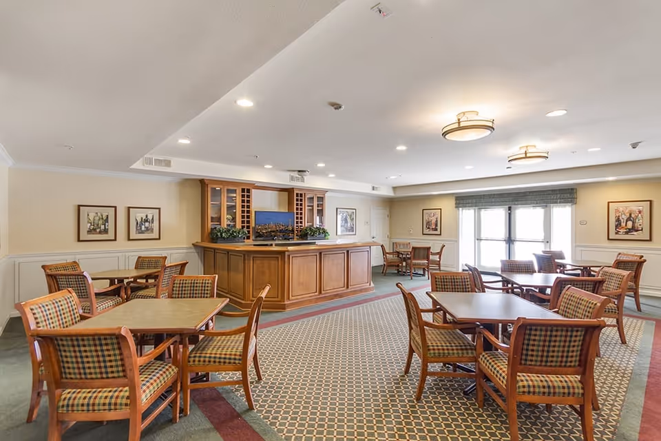 Bright communal dining room with multiple square tables and patterned chairs arranged around a central wooden service counter.
