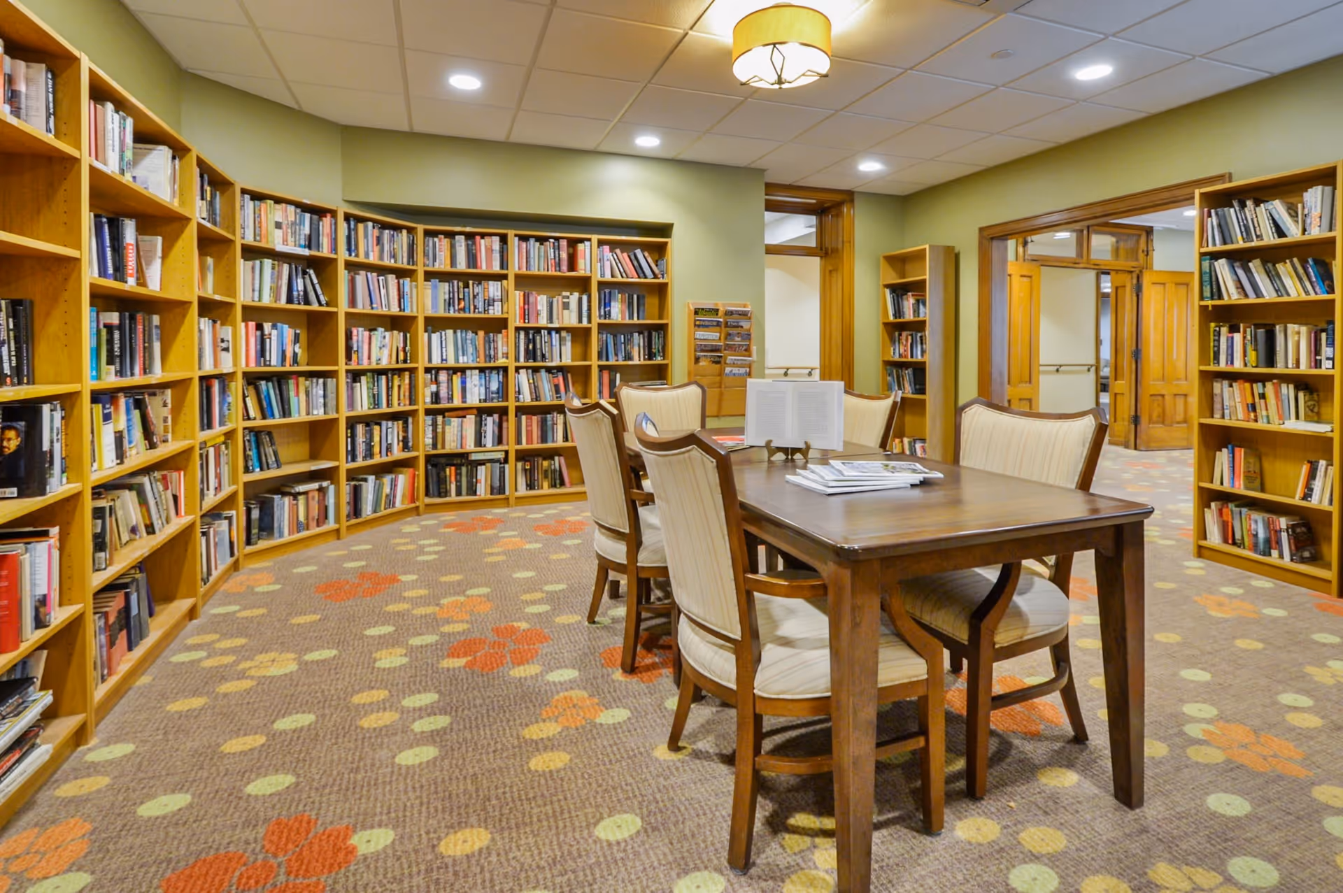 A cozy library room with wooden bookshelves filled with books lining the walls. In the center, there is a wooden table surrounded by six cushioned chairs. The carpet has a floral pattern with orange, yellow, and green colors. The room is well-lit with ceiling lights and a hanging lamp.