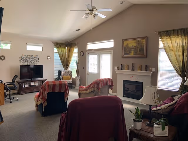 Spacious living room with vaulted ceiling, multiple armchairs covered with throws, a fireplace, TV, and large windows with curtains.