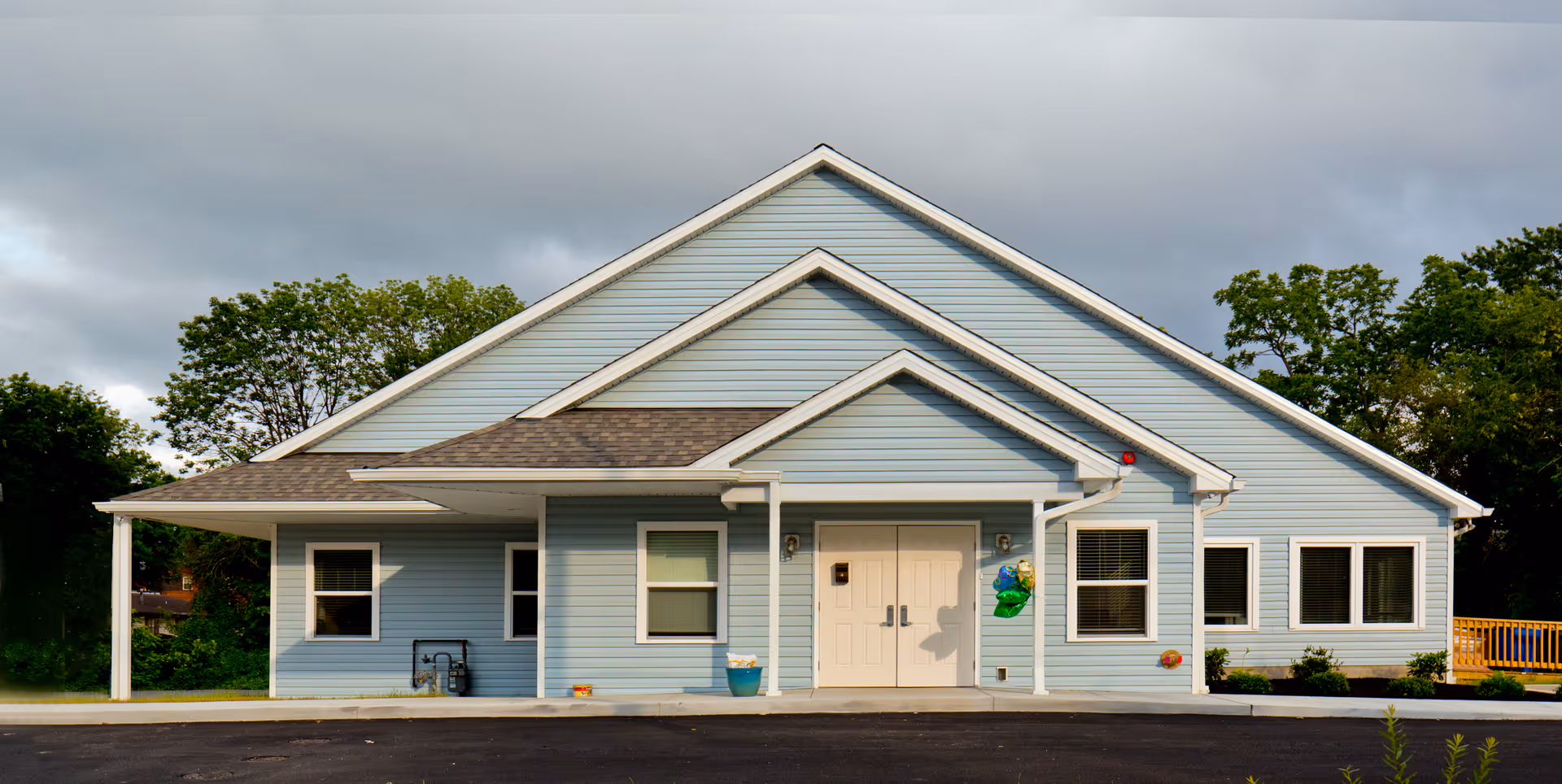 Front exterior view of a light blue building with white trim and a double door entrance, surrounded by greenery and under a cloudy sky.