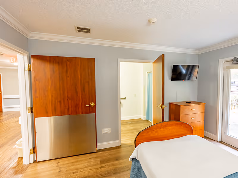 A senior living facility bedroom with a wooden bed partially visible in the foreground, a wooden dresser with a flat-screen TV mounted above it, two wooden doors—one leading to a bathroom with a blue shower curtain, and the other to a hallway. The room has light gray walls, wood flooring, and a glass door leading outside.