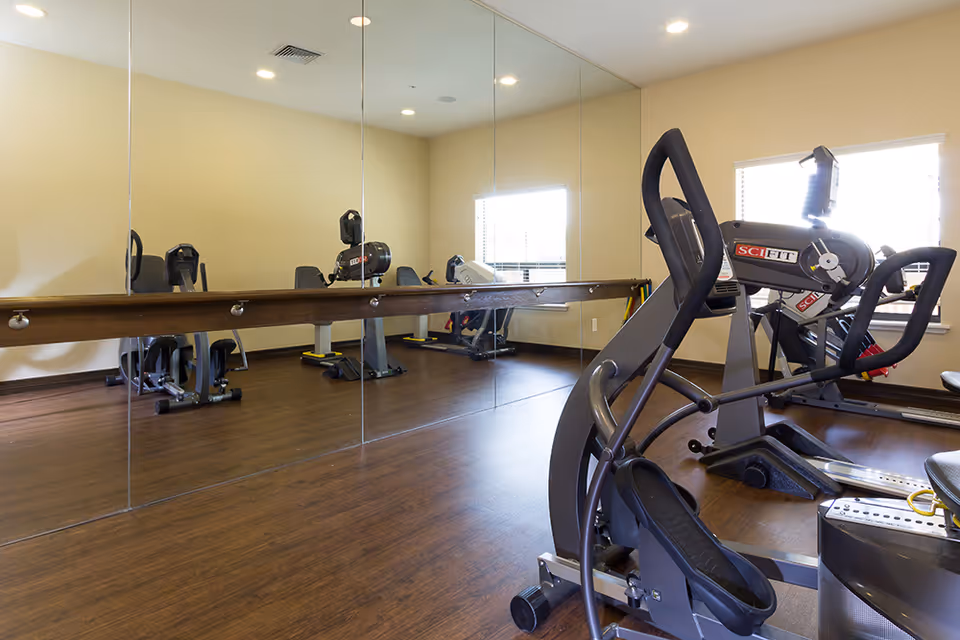 A fitness room with exercise equipment including a SciFit machine, wooden flooring, large wall mirrors, and windows letting in natural light.