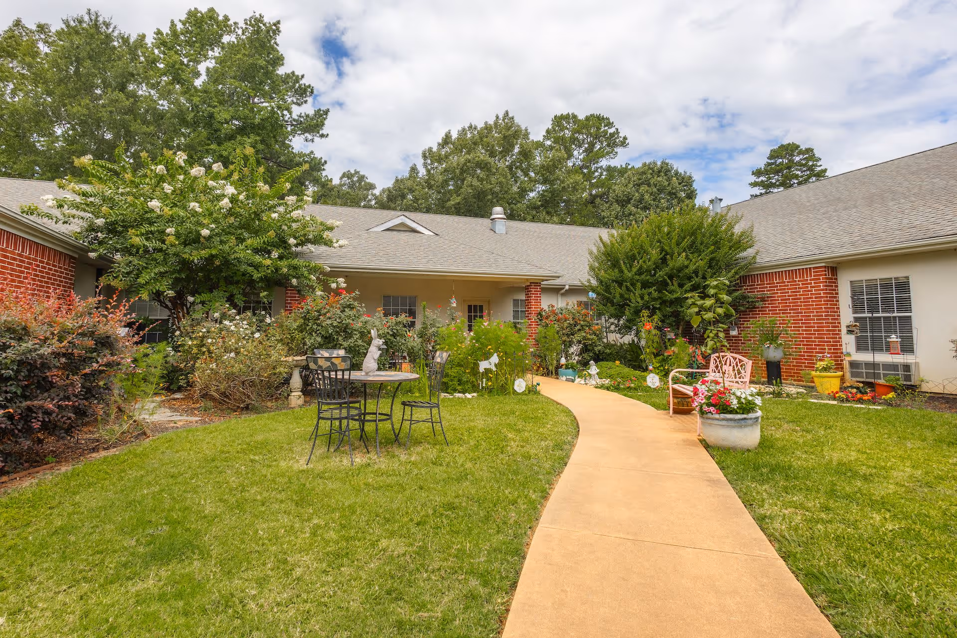 Outdoor garden area at Arabella of Kilgore Senior Living with a paved walkway, green grass, flowering plants, shrubs, a small round table with two chairs, and a pink bench. The building with red brick and white siding is visible in the background under a partly cloudy sky.