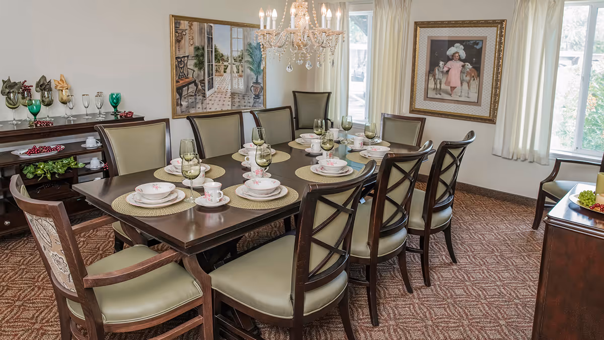 A formal dining room with a dark wooden table set for eight people. The table has beige placemats, floral-patterned dishes, cups, and green-tinted wine glasses. Surrounding the table are cushioned chairs with wooden frames. A chandelier with hanging crystals is above the table. The room has carpeted flooring, two large windows with white curtains, and framed artwork on the walls. A sideboard with decorative glassware and greenery is visible in the background.