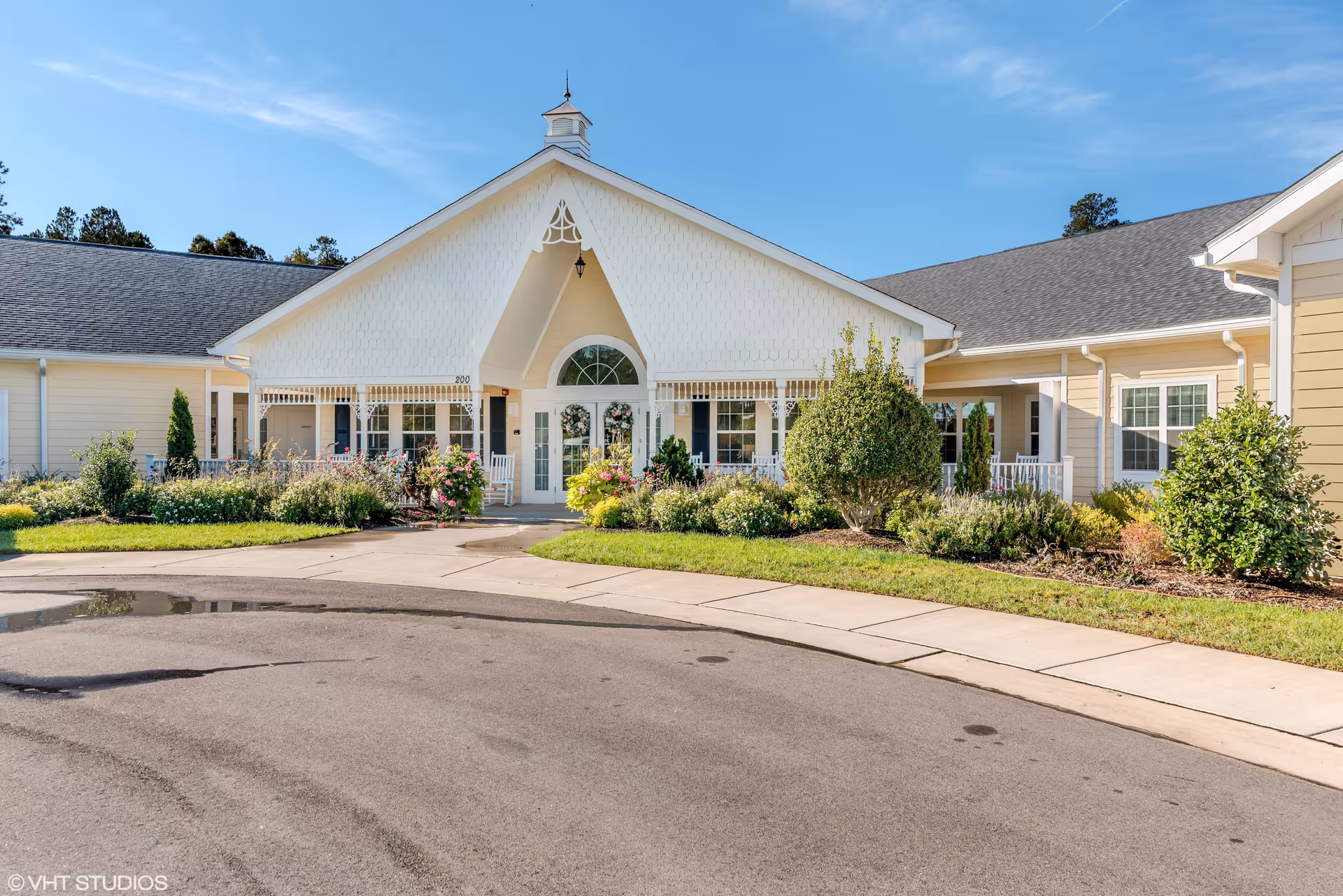 Front exterior view of a single-story building with a peaked roof and white siding. The entrance features double glass doors with decorative wreaths and is flanked by windows and rocking chairs on a porch. There are well-maintained shrubs, flowers, and greenery in front of the building, with a paved driveway and sidewalk leading to the entrance under a clear blue sky.