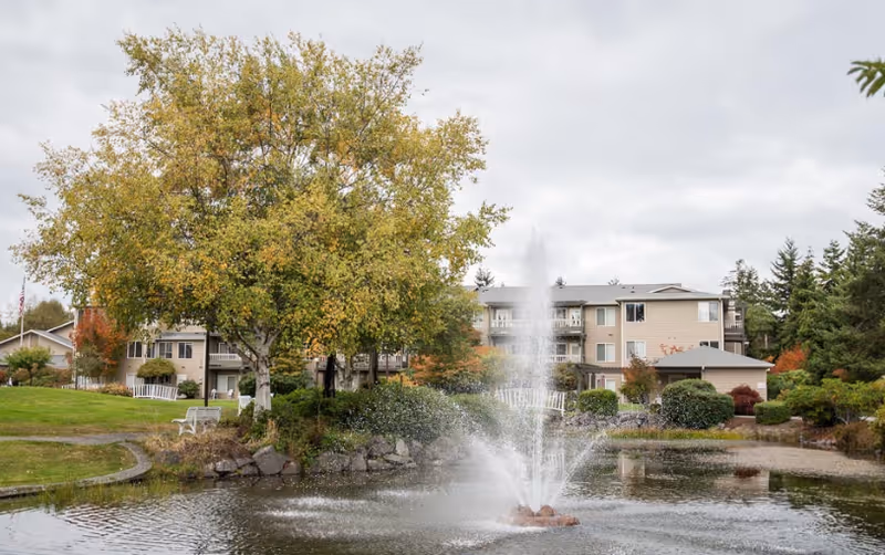 A multi-story senior living building overlooks a pond with a central water fountain and autumn trees.