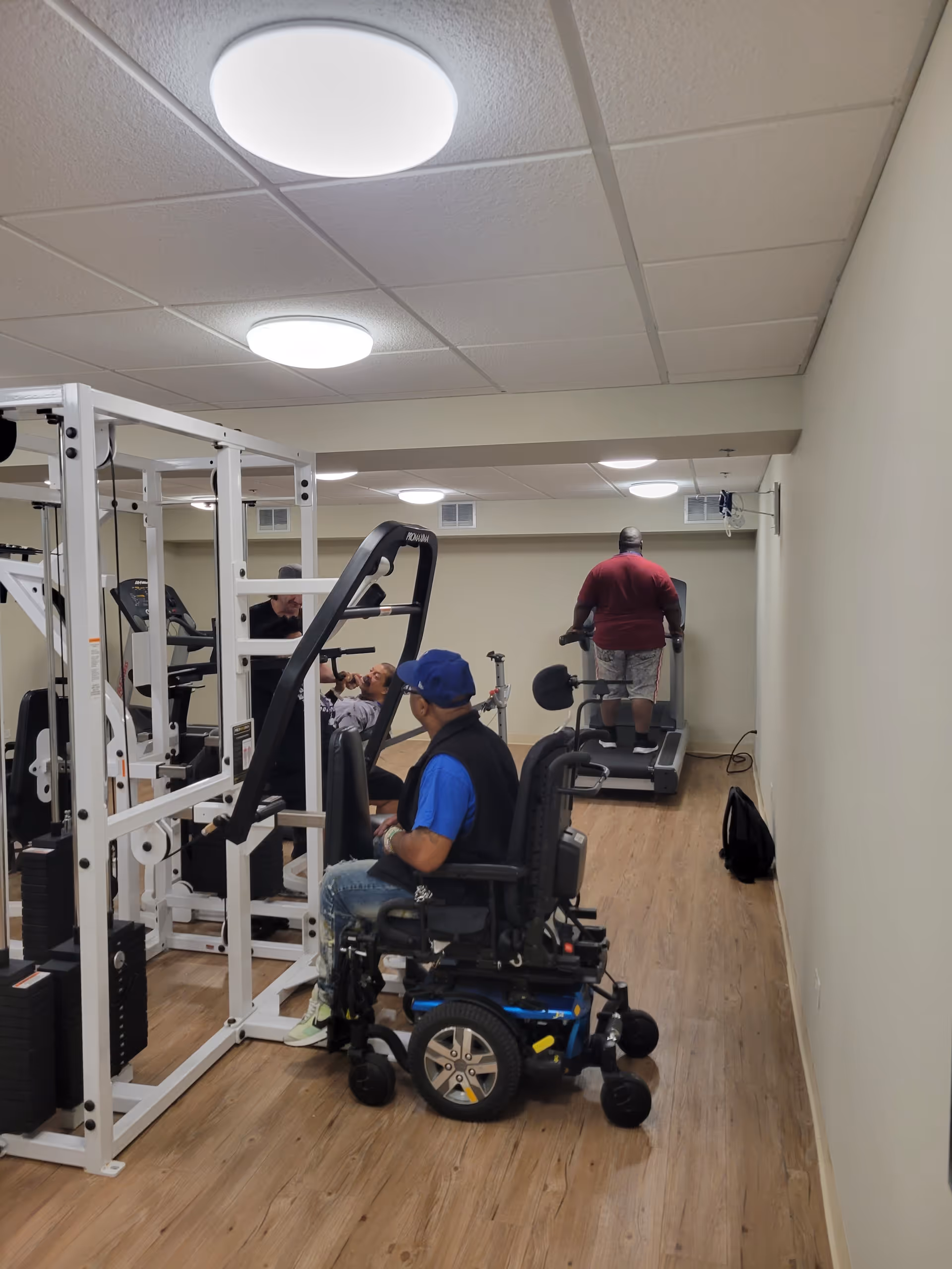 A small fitness room with wooden flooring and beige walls. There are three men using exercise equipment: one man in a wheelchair is seated near a weight machine, another man is lying on a bench lifting weights, and a third man is walking on a treadmill. The room has ceiling lights and a mirror on the left wall.