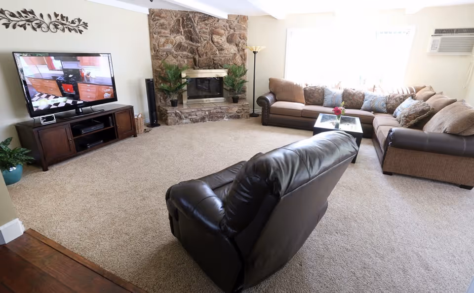 A cozy living room with a large brown sectional sofa adorned with multiple patterned cushions, a dark leather recliner chair, a glass-top coffee table with a small flower vase, a stone fireplace with two potted plants on the hearth, a flat-screen TV on a wooden stand, and a window air conditioning unit.